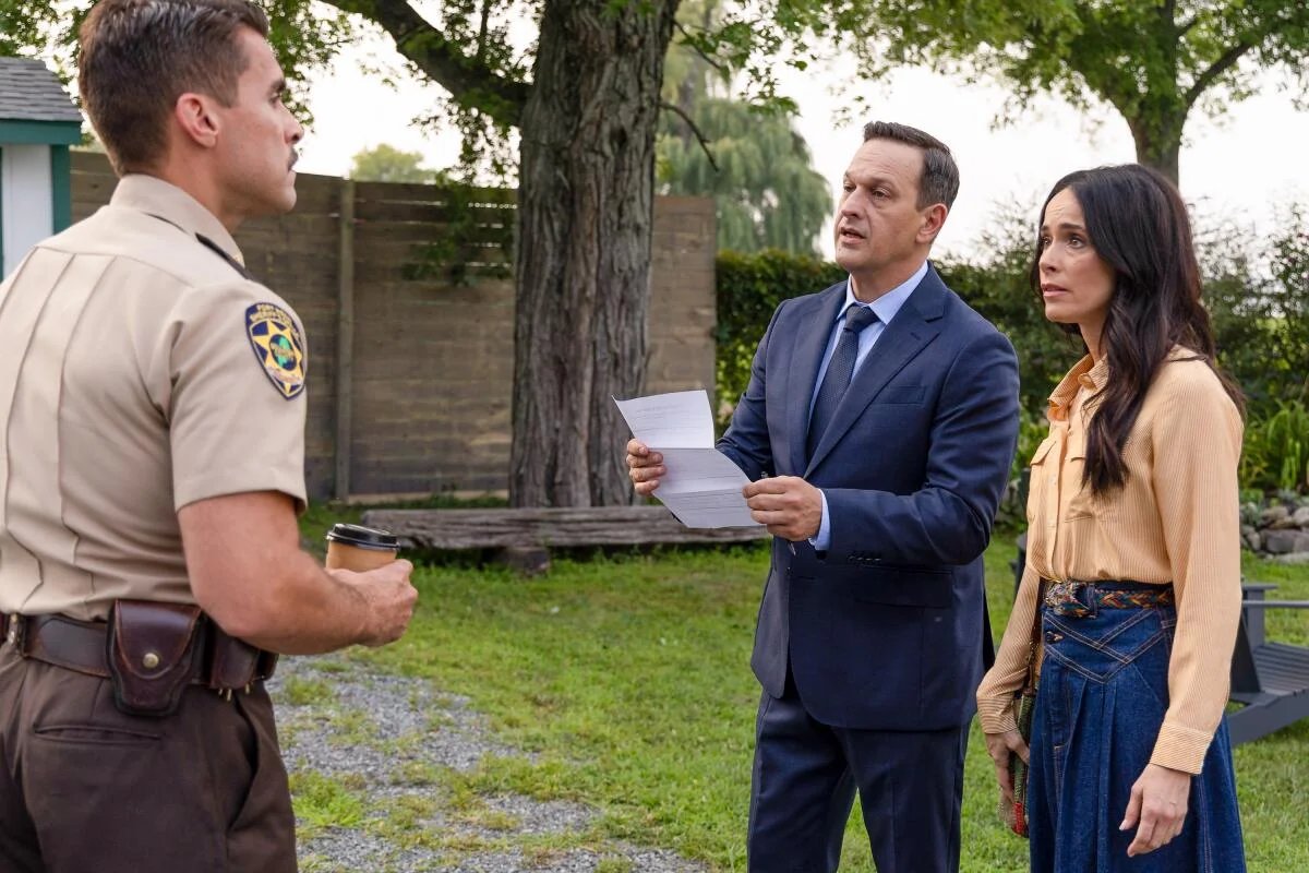 A sheriff in a tan uniform looks at a man in a blue suit and a woman an orange top and denim skirt standing next to him